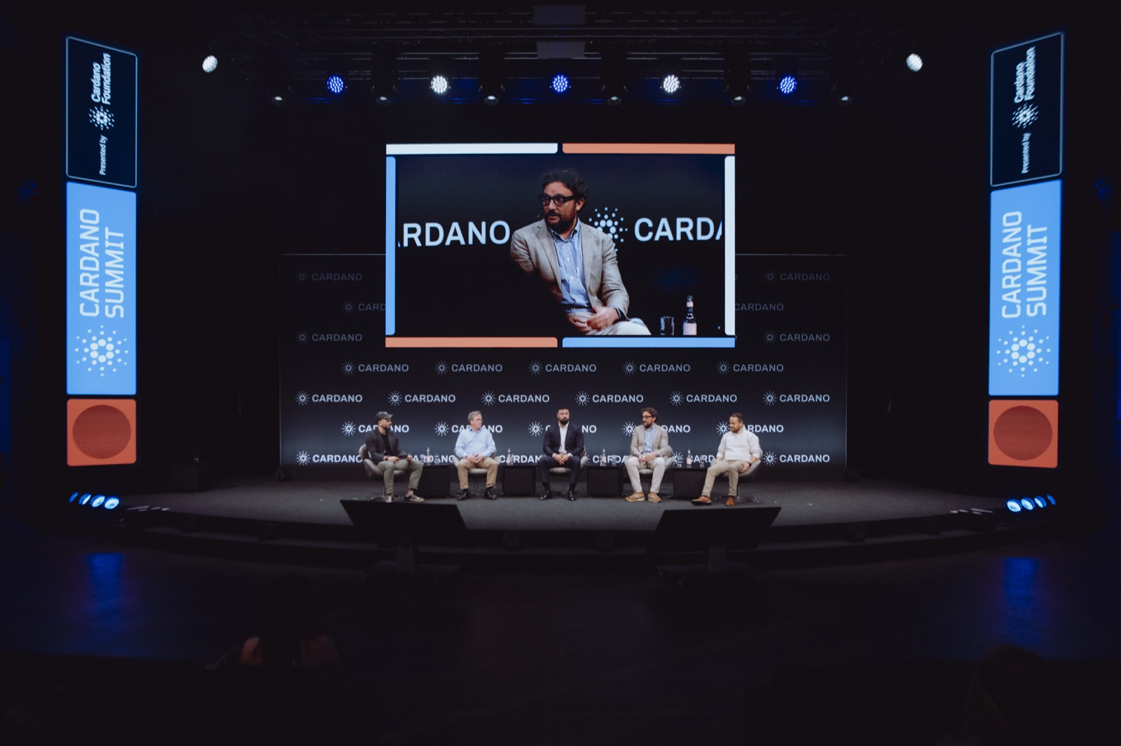 Panel on the Cardano Summit main stage in Berlin, framed by blue and orange brand towers and a large screen